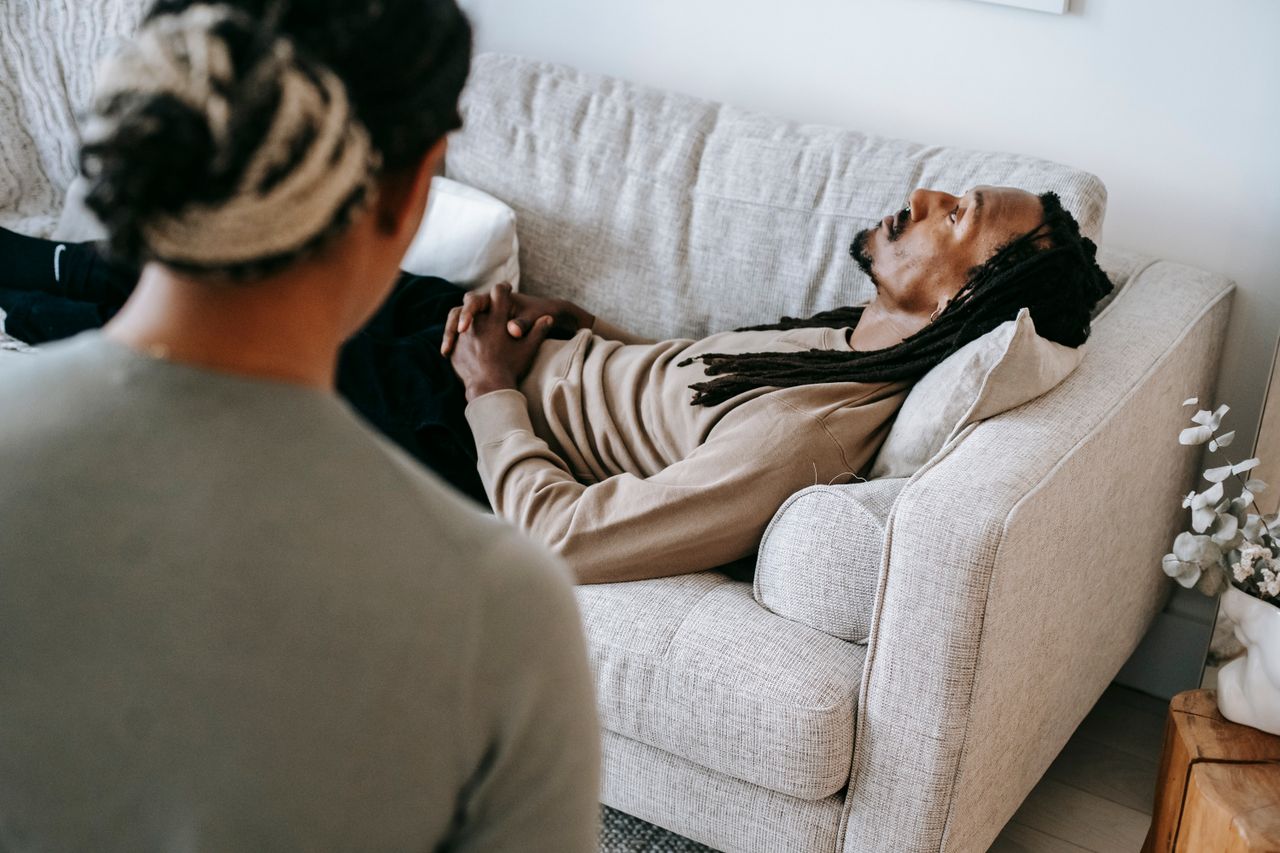 Thoughtful African American male in casual sweater lying on cozy couch and listening to professional female psychologist during psychotherapy